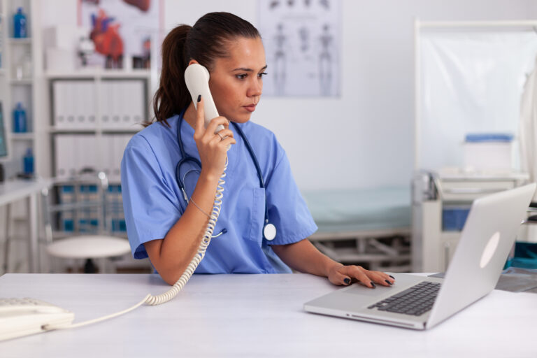 Medical practitioner answering phone calls and scheduling appointments in hospital office. Health care physician sitting at desk using computer in modern clinic looking at monitor.
