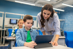 Interesting here. Interested smiling young woman looking into tablet of student boy sitting at desk in school illuminated classroom
