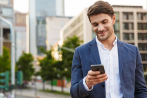 Handsome smiling young businessman smartly dressed standing outdoors at the city street, holding mobile phone